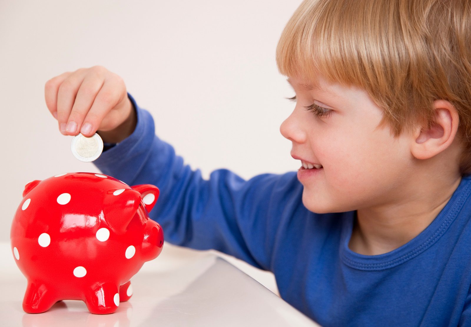Uma criança (menino) colocando uma moeda em um cofrinho vermelho com bolinhas brancas